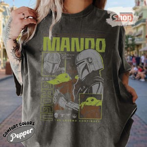 May include: Dark gray t-shirt featuring a graphic of The Mandalorian and Grogu, with the text "MANDO" and "GROGU" in a lime green font. The shirt also includes the text "WHEREVER I GO HE GOES" and "THE LEGEND CONTINUES".