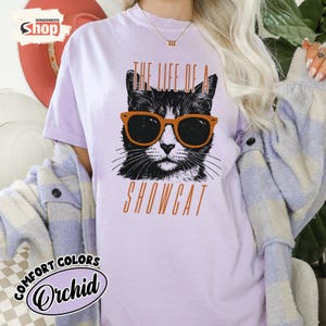May include: Orchid-colored t-shirt with a graphic of a black and white cat wearing orange sunglasses. The text "THE LIFE OF A SHOWCAT" is printed below the cat image in orange. The shirt has the "COMFORT COLORS" logo.