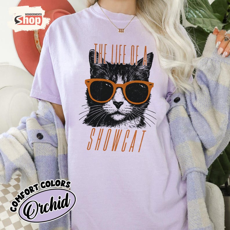 May include: Orchid-colored t-shirt with a graphic of a black and white cat wearing orange sunglasses. The text "THE LIFE OF A SHOWCAT" is printed below the cat image in orange. The shirt has the "COMFORT COLORS" logo.