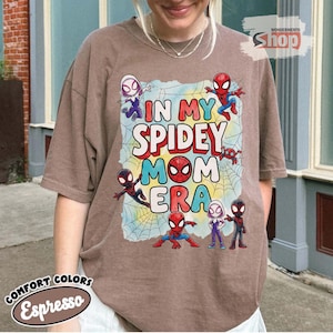 May include: Espresso-coloured t-shirt with the text "IN MY SPIDEY MOM ERA" and Spider-Man characters. The design includes a spider web background and various Spidey characters in action poses. The shirt is from Comfort Colors.