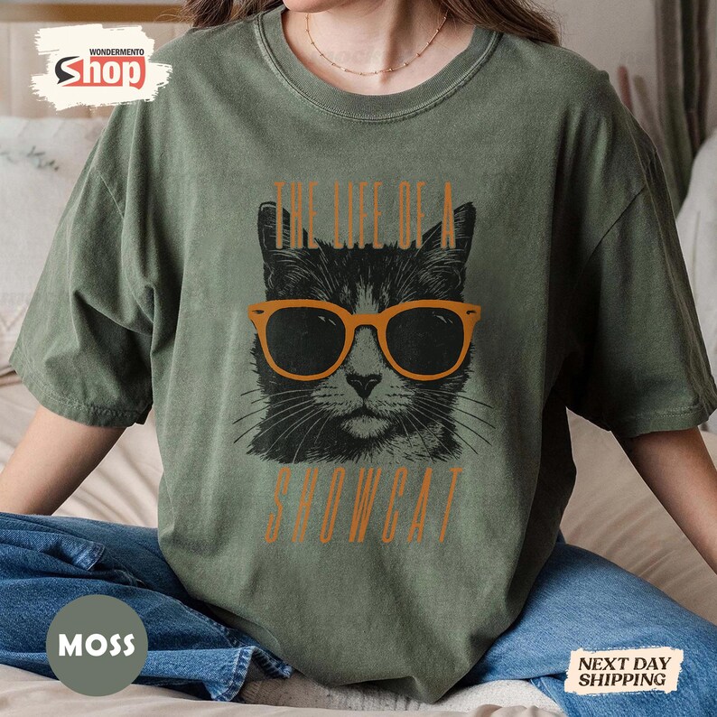 May include: Moss green t-shirt featuring a black cat wearing orange sunglasses. The text "THE LIFE OF A SHOWCAT" is printed below the cat graphic in orange. The shirt has a relaxed fit.