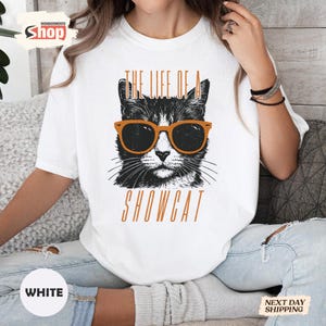 May include: White t-shirt featuring a black and white cat wearing orange sunglasses. The text "THE LIFE OF A SHOWCAT" is printed in orange above and below the cat illustration. The shirt has a crew neck.