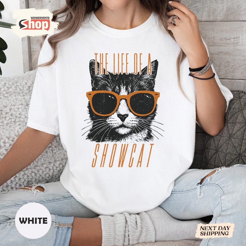 May include: White t-shirt featuring a black and white cat wearing orange sunglasses. The text "THE LIFE OF A SHOWCAT" is printed in orange above and below the cat illustration. The shirt has a crew neck.