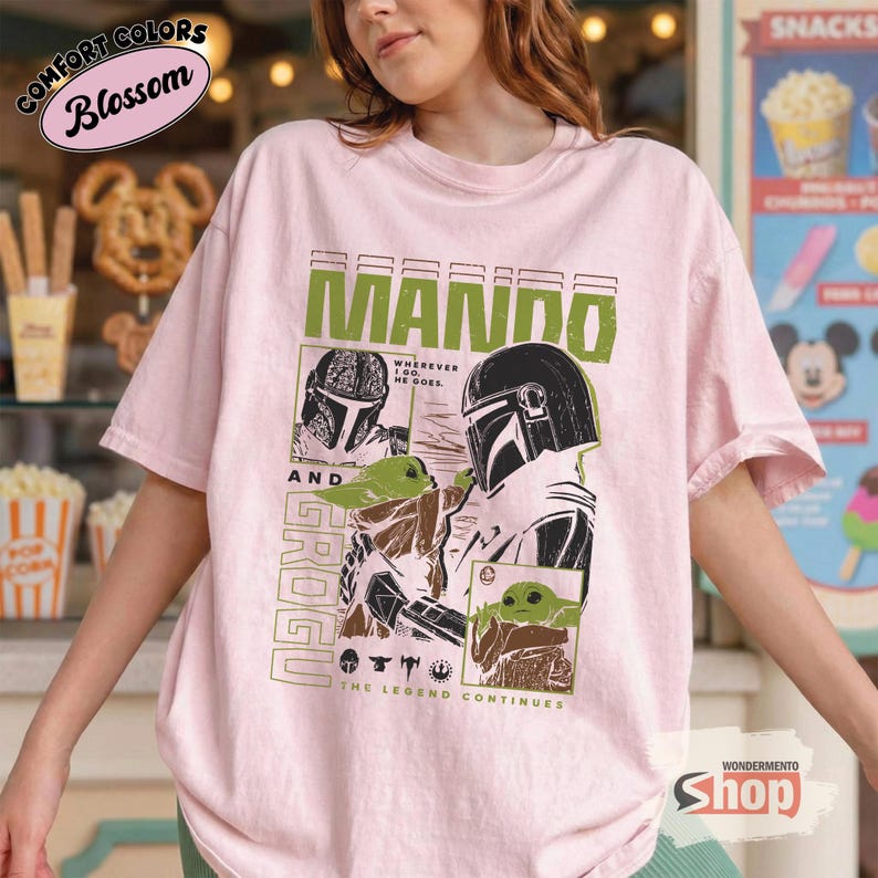 May include: A light pink t-shirt featuring a graphic design with the text "MANDO" in green, and "GROGU" in a vertical font. The design includes illustrations of the Mandalorian and Grogu, with the phrase "THE LEGEND CONTINUES".