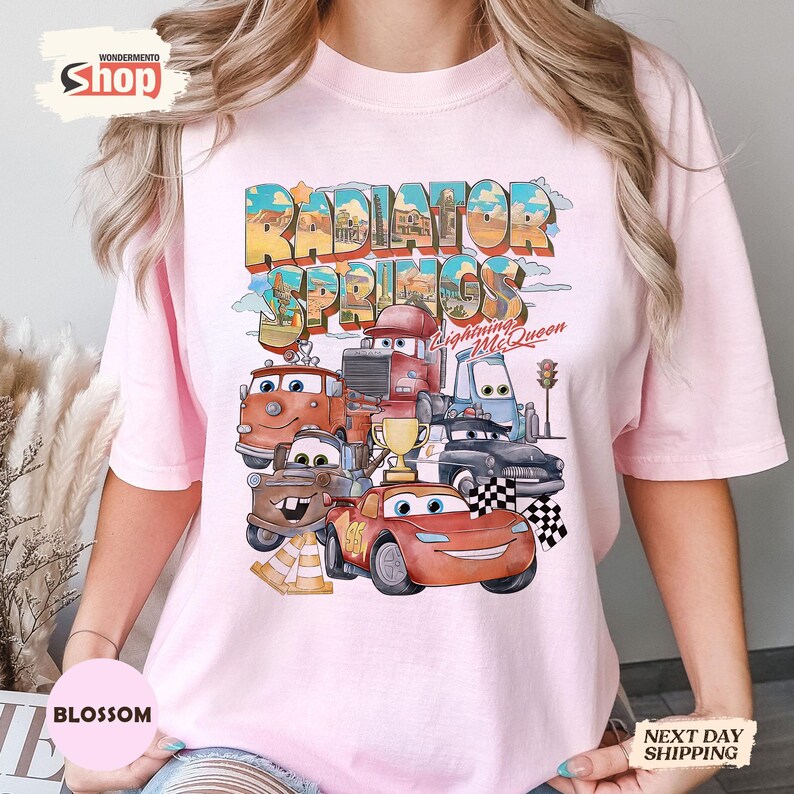 May include: A pale pink t-shirt with a vibrant graphic of characters from the film Cars. The design includes the text "Radiator Springs" and "Lightning McQueen". The graphic features various car characters, a trophy, and a chequered flag.