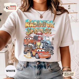 May include: White t-shirt with a colourful graphic of characters from the film Cars, including Lightning McQueen and Mater, and the text "Radiator Springs." The design has a vintage, cartoon style.