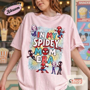 May include: Pale pink t-shirt with the slogan "IN MY SPIDEY MOM ERA" in red, blue, and yellow. The shirt features cartoon images of Spider-Man characters and spiderwebs. The shirt has the "Comfort Colors" and "Blossom" logo.