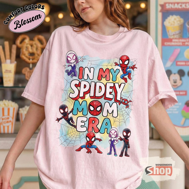 May include: Pale pink t-shirt with the slogan "IN MY SPIDEY MOM ERA" in red, blue, and yellow. The shirt features cartoon images of Spider-Man characters and spiderwebs. The shirt has the "Comfort Colors" and "Blossom" logo.