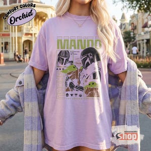 May include: Orchid-colored t-shirt featuring a graphic of Mando and Grogu from The Mandalorian. The design includes the text "MANDO" in green block letters, and "GROGU" vertically. The shirt also has the text "Wherever he goes. The legend continues."