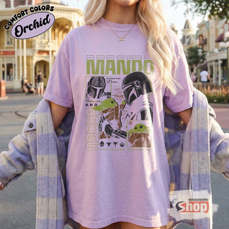 May include: Orchid-colored t-shirt featuring a graphic of Mando and Grogu from The Mandalorian. The design includes the text "MANDO" in green block letters, and "GROGU" vertically. The shirt also has the text "Wherever he goes. The legend continues."