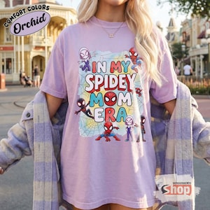 May include: A lilac t-shirt featuring the phrase "IN MY SPIDEY MOM ERA" with cartoon Spider-Man characters. The shirt is from the "WONDERMENTO SHOP" and is part of the "Comfort Colors" collection.