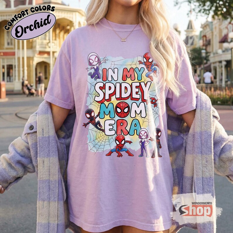 May include: A lilac t-shirt featuring the phrase "IN MY SPIDEY MOM ERA" with cartoon Spider-Man characters. The shirt is from the "WONDERMENTO SHOP" and is part of the "Comfort Colors" collection.