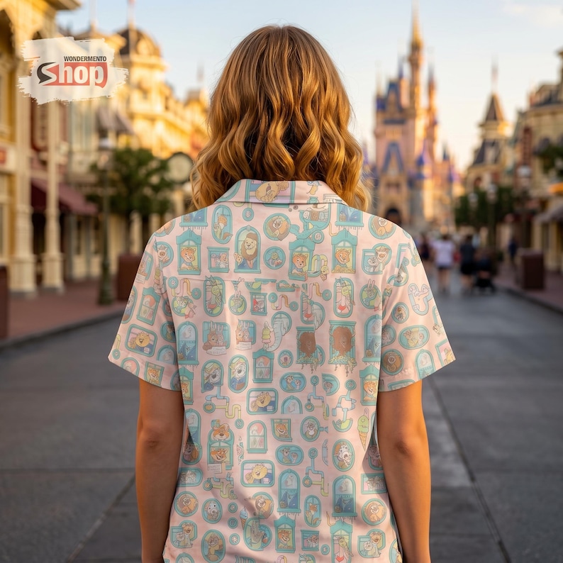 May include: A light pink button-up shirt with a repeating pattern of cartoon characters in ornate frames. The shirt has short sleeves and a collared neckline. The background shows a theme park with a castle.