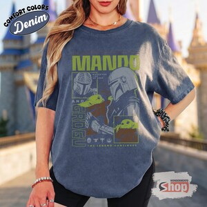 May include: Denim blue t-shirt featuring a graphic of Mando and Grogu from The Mandalorian. The design includes the text "Mando and Grogu" in green, along with images of the characters. The shirt also has the text "The Legend Continues".