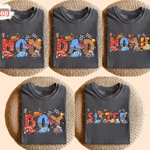 May include: Five dark gray t-shirts with colorful text and car graphics. The shirts read "MOM", "DAD", "NOAH", "BOY", and "SISTER". Each shirt features a checkered flag and the number "95".