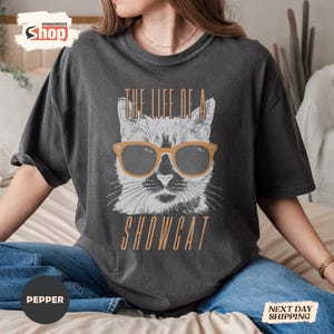 May include: Dark gray t-shirt featuring a cat wearing sunglasses with the text "THE LIFE OF A SHOWCAT" in a retro font. The cat graphic is white with a tan outline. The shirt is a relaxed fit.
