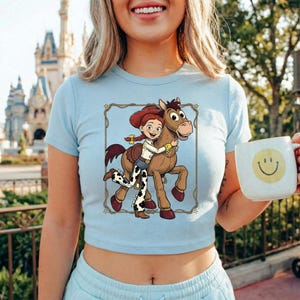 Peut inclure: Haut court bleu clair avec un dessin anim&eacute; d'une fille &eacute;treignant un cheval. Le dessin est encadr&eacute; d'une bordure d&eacute;corative. Une tasse blanche avec un visage souriant jaune est tenue dans la main.