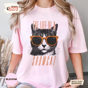 May include: A light pink t-shirt featuring a black and white cat wearing orange sunglasses. The text "THE LIFE OF A SHOWCAT" is printed below the cat image in orange letters. The shirt is a casual style.