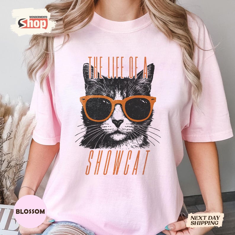 May include: A light pink t-shirt featuring a black and white cat wearing orange sunglasses. The text "THE LIFE OF A SHOWCAT" is printed below the cat image in orange letters. The shirt is a casual style.