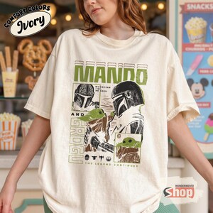 May include: Ivory-colored t-shirt featuring a graphic of The Mandalorian and Grogu. The design includes the text "MANDO AND GROGU" in green, along with illustrations of the characters. The shirt also has the text "THE LEGEND CONTINUES".