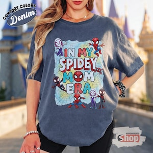 May include: A denim blue t-shirt with the text "IN MY SPIDEY MOM ERA" in colourful letters and Spider-Man characters. The shirt has a distressed look and features the words "COMFORT COLORS" and "Denim" in a circular design.