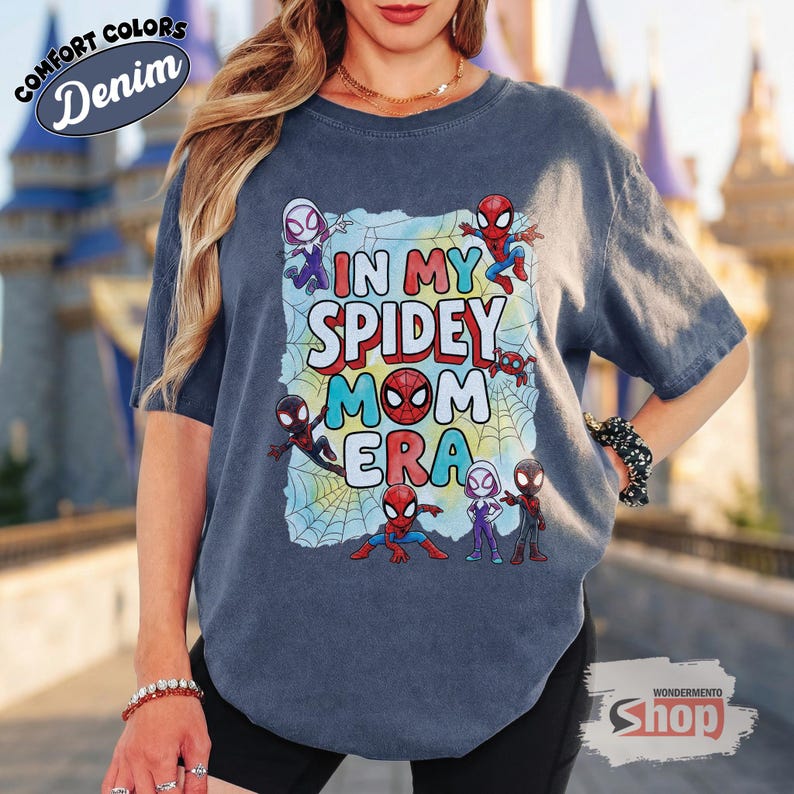 May include: A denim blue t-shirt with the text "IN MY SPIDEY MOM ERA" in colourful letters and Spider-Man characters. The shirt has a distressed look and features the words "COMFORT COLORS" and "Denim" in a circular design.
