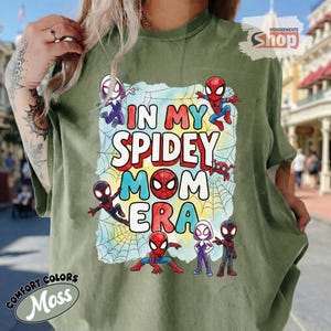 May include: A moss green t-shirt with the words "IN MY SPIDEY MOM ERA" in a colourful design. The graphic includes Spider-Man characters and a spiderweb. The shirt is from Comfort Colors.