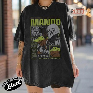 May include: Black oversized t-shirt featuring a graphic design with the text "MANDO AND GROGU" in green and white. The design includes illustrations of the Mandalorian and Grogu. The shirt also has the text "THE LEGEND CONTINUES".