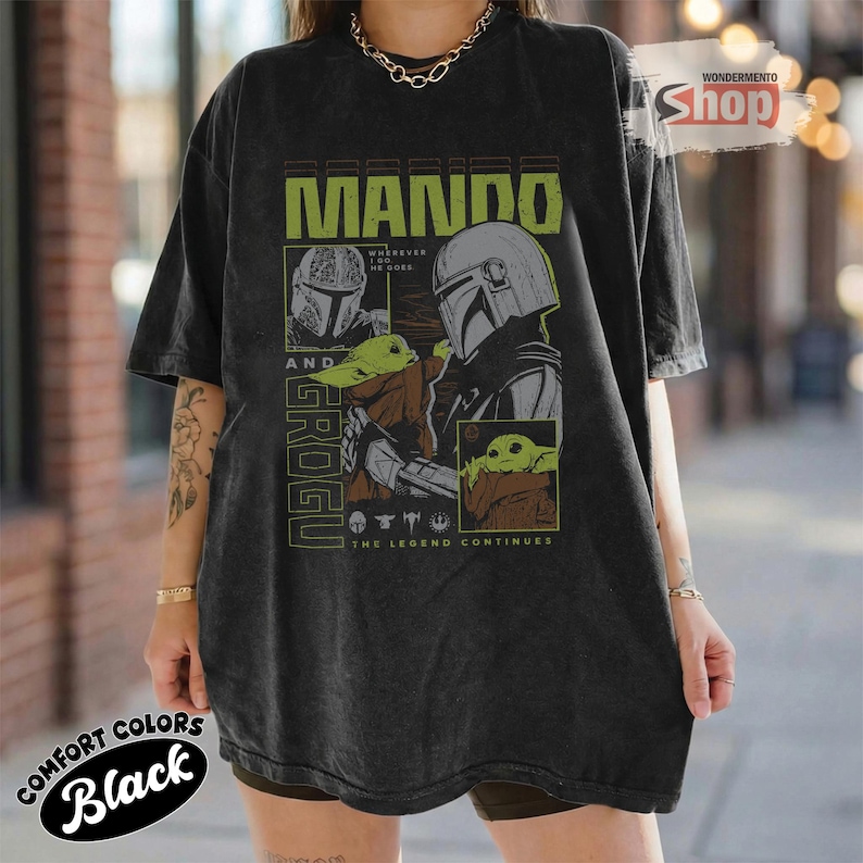 May include: Black oversized t-shirt featuring a graphic design with the text "MANDO AND GROGU" in green and white. The design includes illustrations of the Mandalorian and Grogu. The shirt also has the text "THE LEGEND CONTINUES".