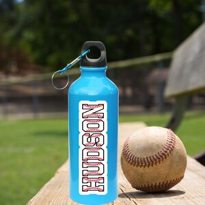 May include: A blue water bottle with a black lid and a white baseball-themed sticker with the name "HUDSON" on it. The bottle is sitting on a wooden surface next to a baseball.