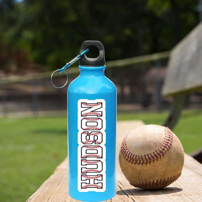 May include: A blue water bottle with a black lid and a white baseball-themed sticker with the name "HUDSON" on it. The bottle is sitting on a wooden surface next to a baseball.