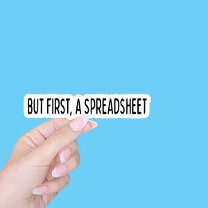 May include: A white sticker with the text "BUT FIRST, A SPREADSHEET" on a light blue background. A hand is holding the sticker.