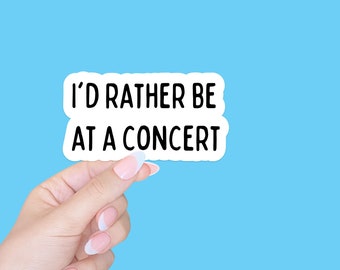 May include: White sticker with black text that reads "I'd rather be at a concert". A hand is holding the sticker against a light blue background.