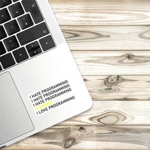 May include: A silver laptop with a sticker that reads "I hate programming I hate programming I hate programming I love programming". The laptop is on a light brown wooden surface.
