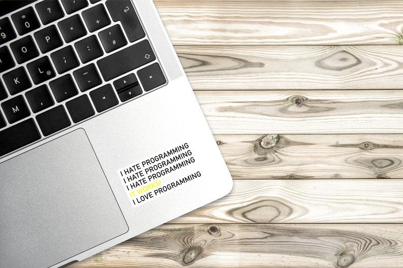 May include: A silver laptop with a sticker that reads "I hate programming I hate programming I hate programming I love programming". The laptop is on a light brown wooden surface.