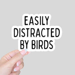 Może przedstawiać: Biała naklejka z napisem "EASILY DISTRACTED BY BIRDS" pogrubioną czcionką. Naklejka trzymana jest przez dłoń z zadbanymi paznokciami na szarym tle. Zabawny i oryginalny przedmiot.