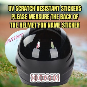 May include: Black baseball helmet with a white baseball next to it. The helmet has a red and white baseball stitch design on the back with the name "HUDSON" in white letters.