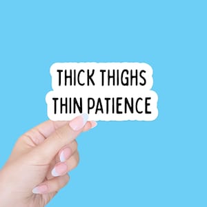Może przedstawiać: Biała naklejka z czarnym napisem "THICK THIGHS THIN PATIENCE", trzymana przez dłoń z zadbanymi paznokciami na jasnoniebieskim tle. Naklejka ma białą obwódkę.