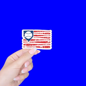 May include: A hand holding a sticker of a distressed American flag with a baseball in the top left corner. The baseball is inside a blue baseball diamond.