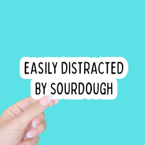 May include: A white sticker with the text "EASILY DISTRACTED BY SOURDOUGH" in black, held by a hand with manicured nails. The sticker has a rounded edge and is set against a bright turquoise background.
