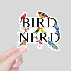 May include: A sticker featuring the words "BIRD NERD" in bold black letters, surrounded by watercolor illustrations of various colorful birds, including a cardinal, hummingbird, and peacock. The sticker is held by a hand.