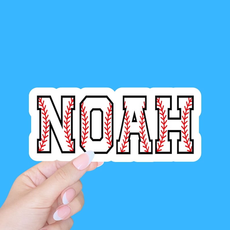 May include: White sticker with the name NOAH in black letters with red stitching that looks like a baseball. The sticker is being held by a hand.