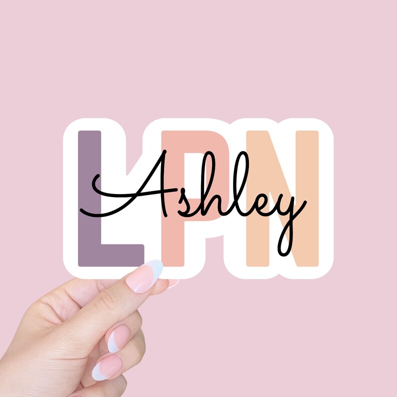 May include: A sticker with the letters LPN in pastel colors, with the name Ashley written in cursive in black. The sticker is being held by a hand with white nail polish.