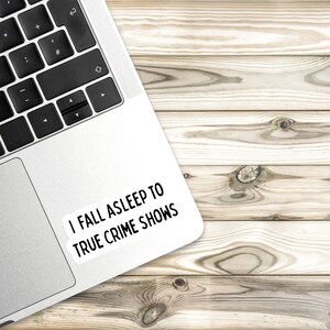 May include: A silver laptop computer with a white sticker on the top right corner that says "I fall asleep to true crime shows". The laptop is on a light brown wooden surface.