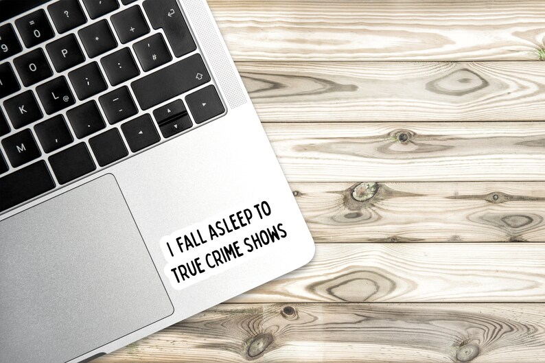 May include: A silver laptop computer with a white sticker on the top right corner that says "I fall asleep to true crime shows". The laptop is on a light brown wooden surface.