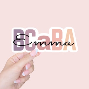 May include: A sticker with the text "BCaBA" in pink, orange, and purple with the word "Emma" written in black cursive underneath.