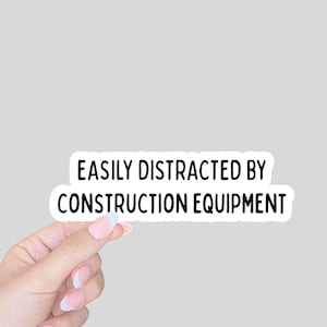 May include: A white sticker with black text that reads "EASILY DISTRACTED BY CONSTRUCTION EQUIPMENT." The sticker is held by a hand with manicured nails against a gray background.