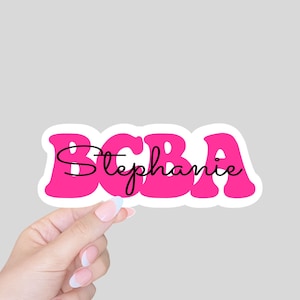 May include: Pink sticker with the letters "BCBA" in a retro font. The name "Stephanie" is written in cursive inside the letters.