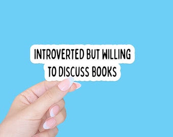 May include: A white sticker with black text that reads "Introverted but willing to discuss books". A hand is holding the sticker against a light blue background.