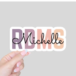 May include: A sticker with the name "Michelle" in a cursive font, overlaying the letters "RDMS" in a block font. The letters are in shades of purple, pink, and peach. The sticker is held by a hand with a light gray background.